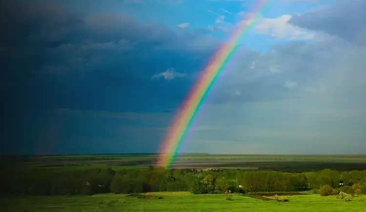 A vibrant rainbow emerges from a dark, stormy sky, casting light over a peaceful green landscape, symbolizing the hope and healing that can follow a period of distress.